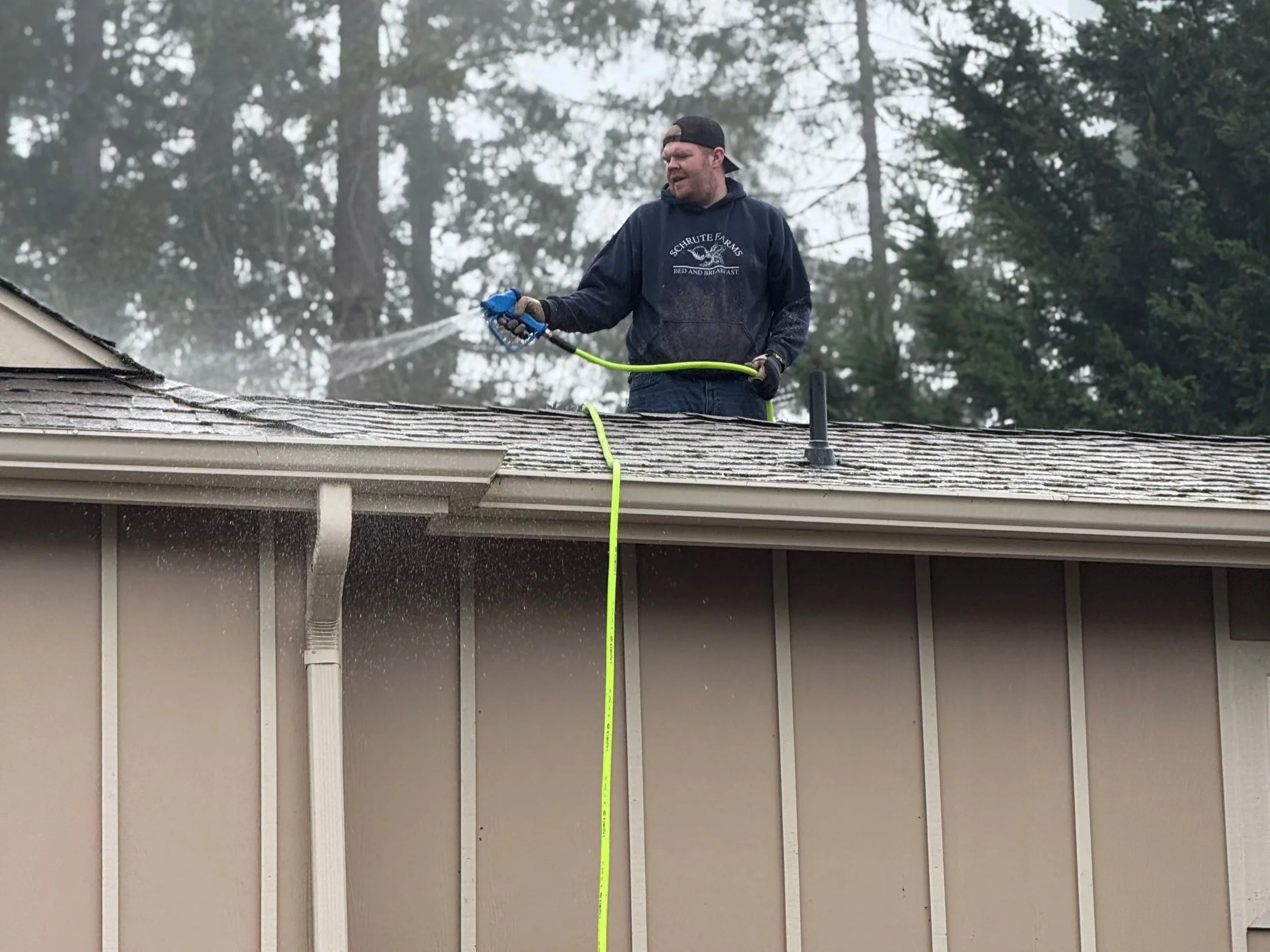 A man standing on a roof cleaning it with a hose, water spray visible.