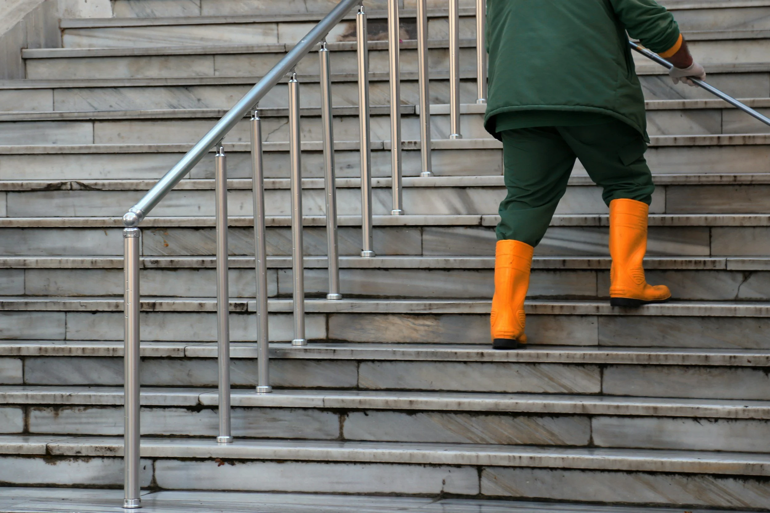 A person wearing orange rubber boots and green clothing is ascending outdoor stone stairs with a metal handrail.