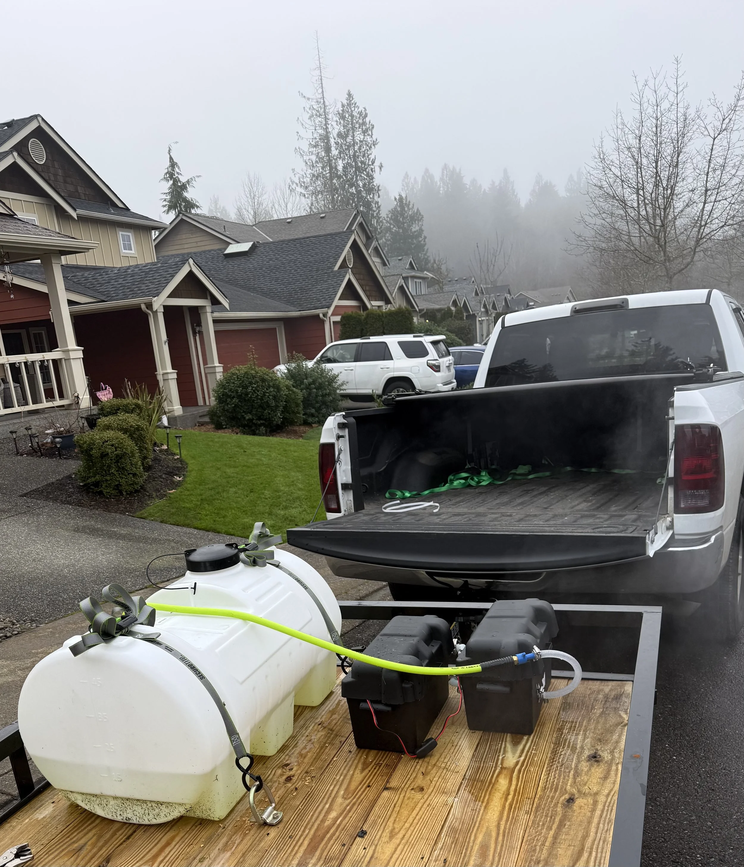 A white pickup truck with its tailgate down, connected to a large white tank and equipment on a wooden platform, parked on a residential street with houses, cars, and trees in the background.