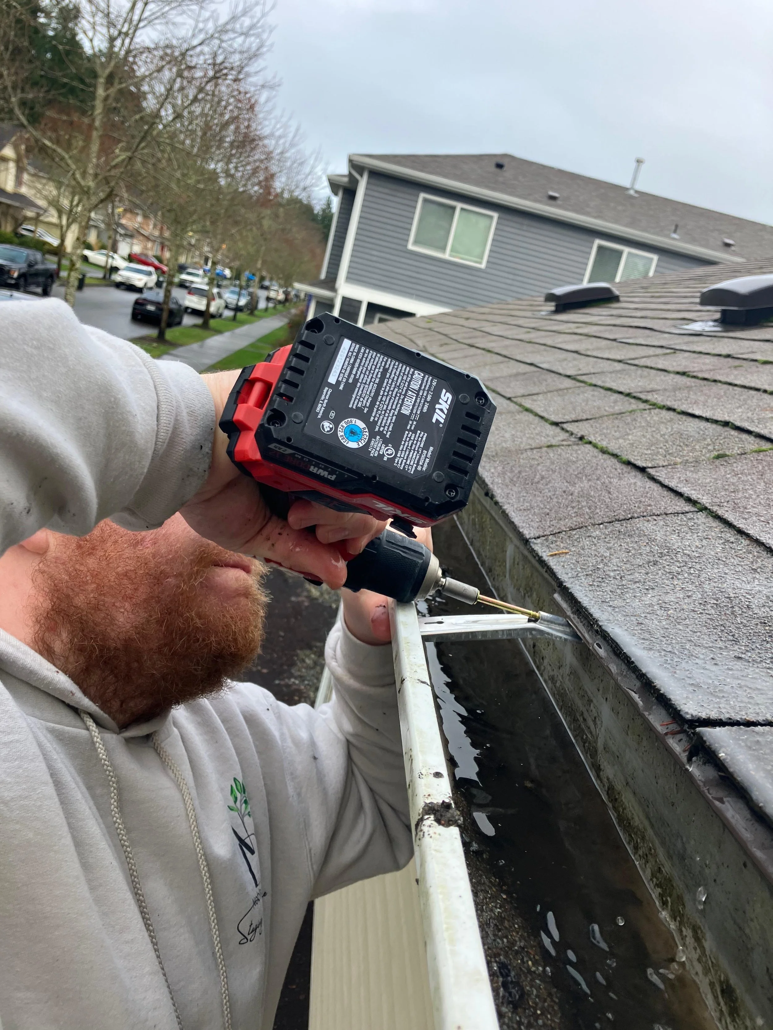 A man fixing or inspecting the gutter of a house with a power drill. The house has asphalt shingles, and in the background, there are other houses, trees, and cars parked on the street.
