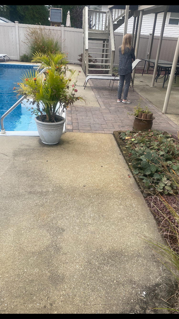 A backyard scene with a swimming pool on the left, potted plants, a woman standing on a brick patio holding a book, wooden stairs leading up to a deck, patio furniture, and a white fence in the background.