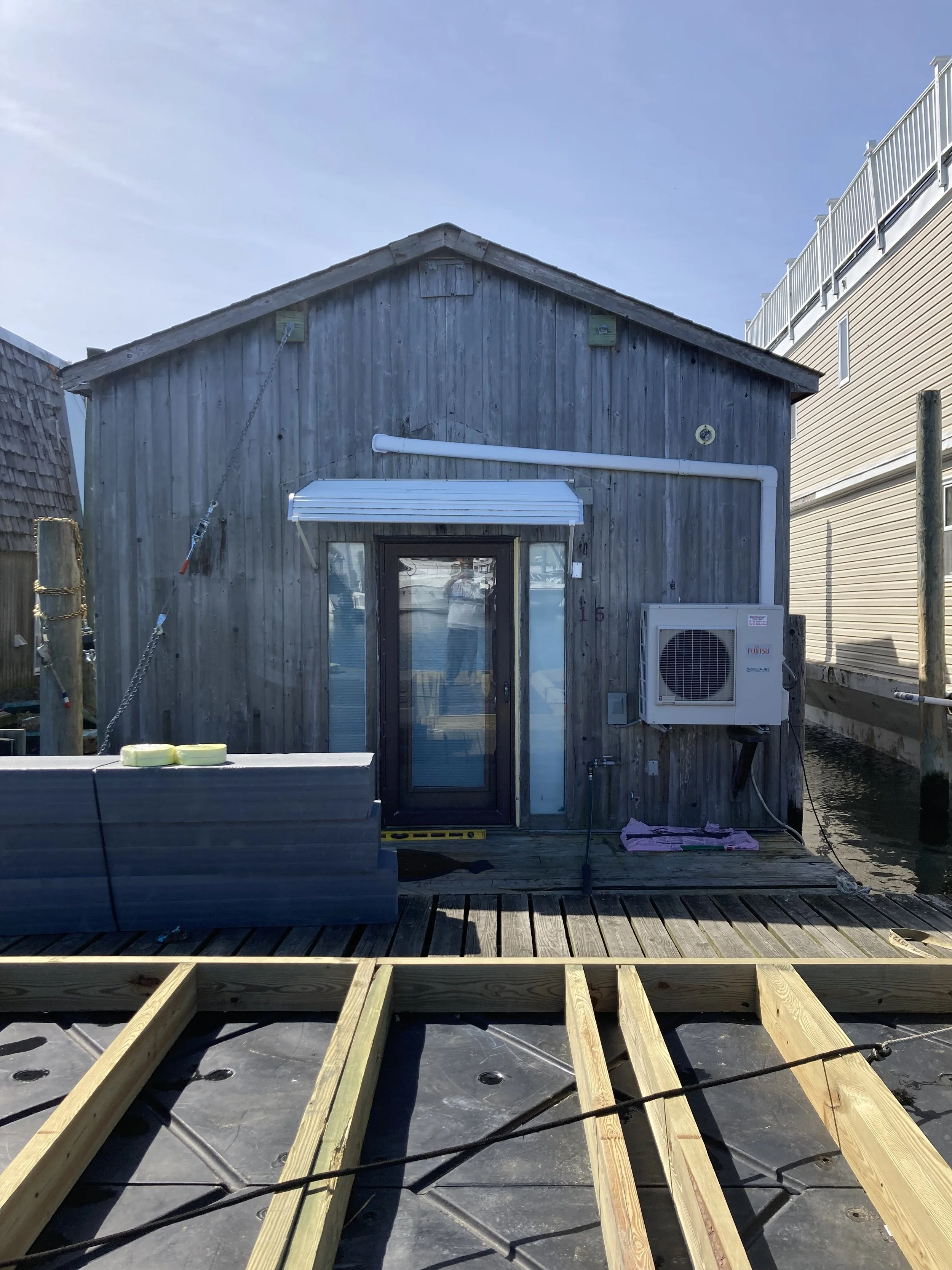 A small wooden house by the water under a clear blue sky with construction materials and partially built deck in the foreground.