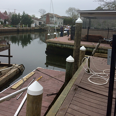 People gathered on a wooden dock or pier beside a canal, with some working or inspecting equipment near the water's edge, and houses or buildings in the background.