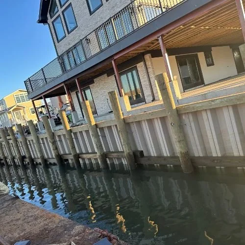 A waterfront house with a balcony overlooking a canal, supported by wooden pilings, with a concrete seawall and clear blue sky.