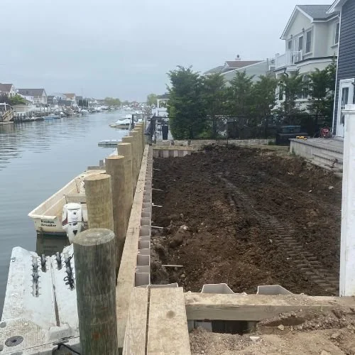 A construction site next to a canal with residential houses. The site has freshly dug soil inside a partially built retaining wall, with some concrete blocks stacked nearby. Several boats are docked along the canal, and there are overcast skies.