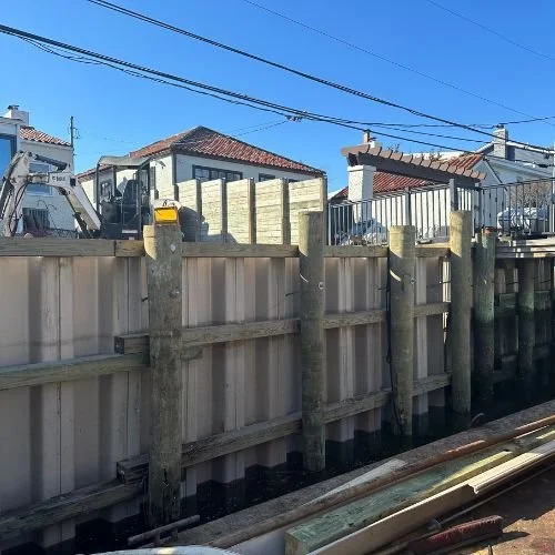 View of a construction site next to a body of water with a wooden and metal retaining wall, a small excavator, and residential houses in the background under a clear blue sky.