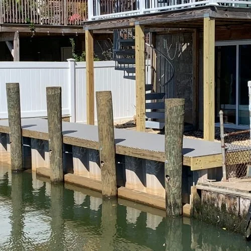 Newly built dock with wooden pilings, concrete walkway, and a spiral staircase leading to an upper deck, located by a residential backyard with a white fence.