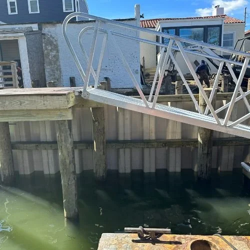 A metal gangway leading from a dock to a house with a deck, over water, with visible support posts in the water.