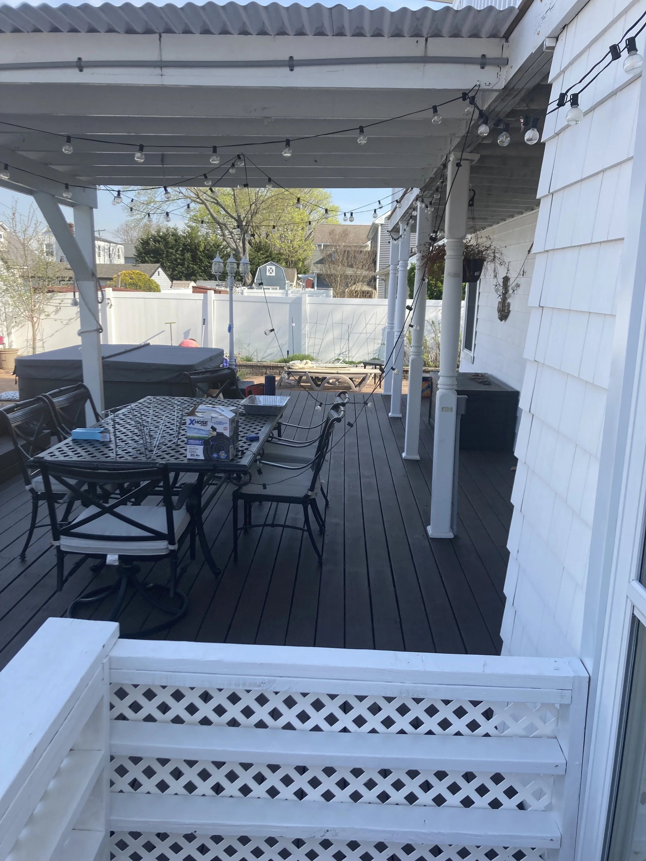 View of a backyard patio with string lights, outdoor furniture including a table and chairs, a hot tub, and garden area enclosed by a white fence.