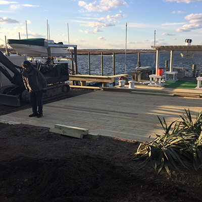 Person working on a wooden dock by the water, with boats and pilings in the background, under a partly cloudy sky.