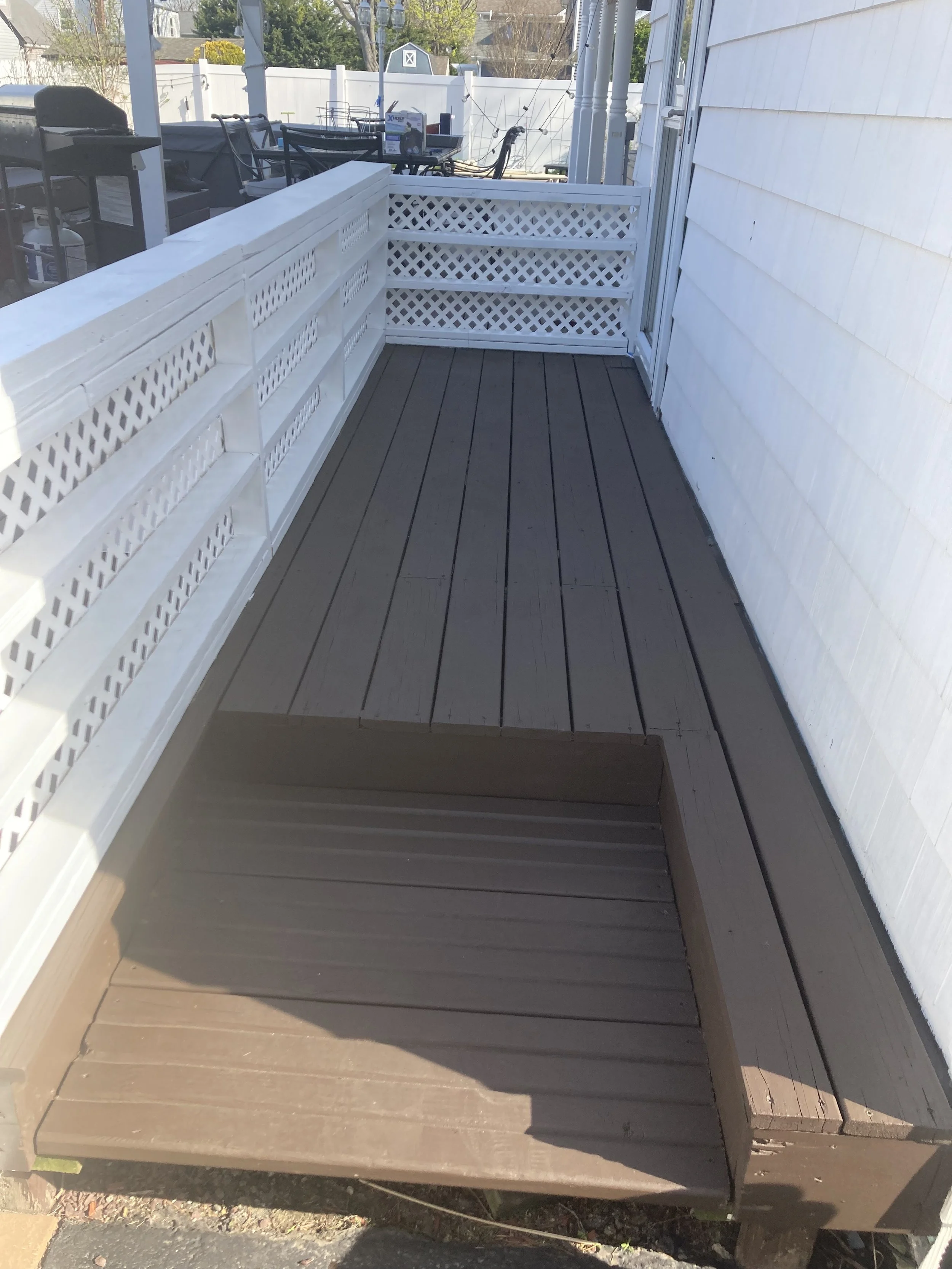 Backyard patio with wooden decking, white lattice fencing, and outdoor furniture including a grill and a table with chairs.