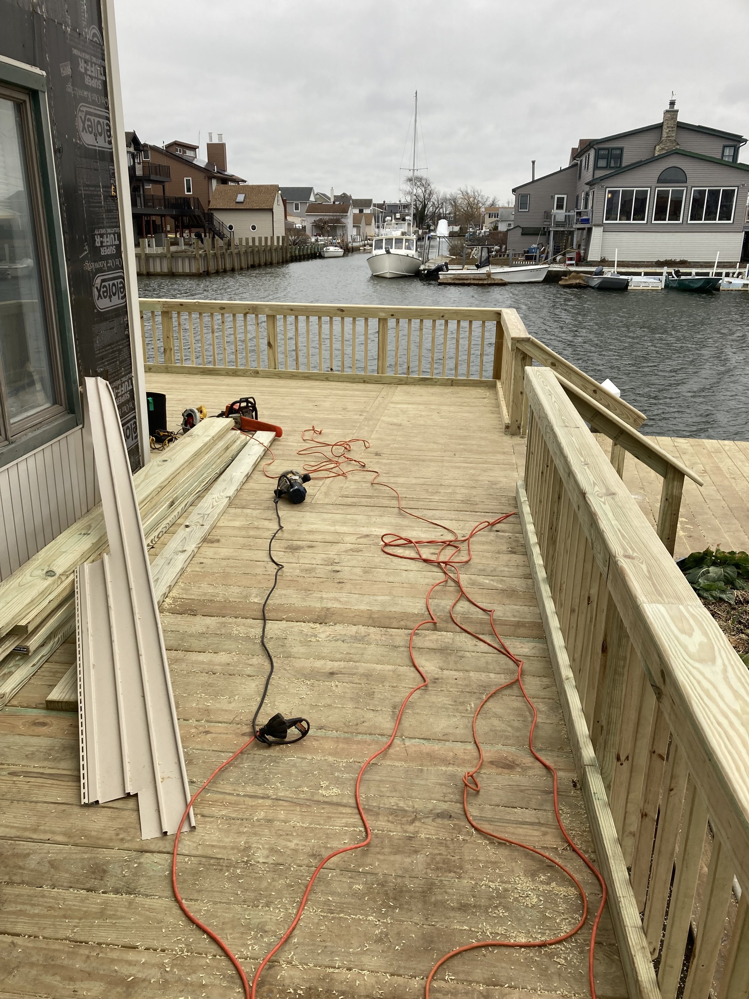 View of a wooden deck under construction, with tools and materials, overlooking a canal with boats and houses on a cloudy day.