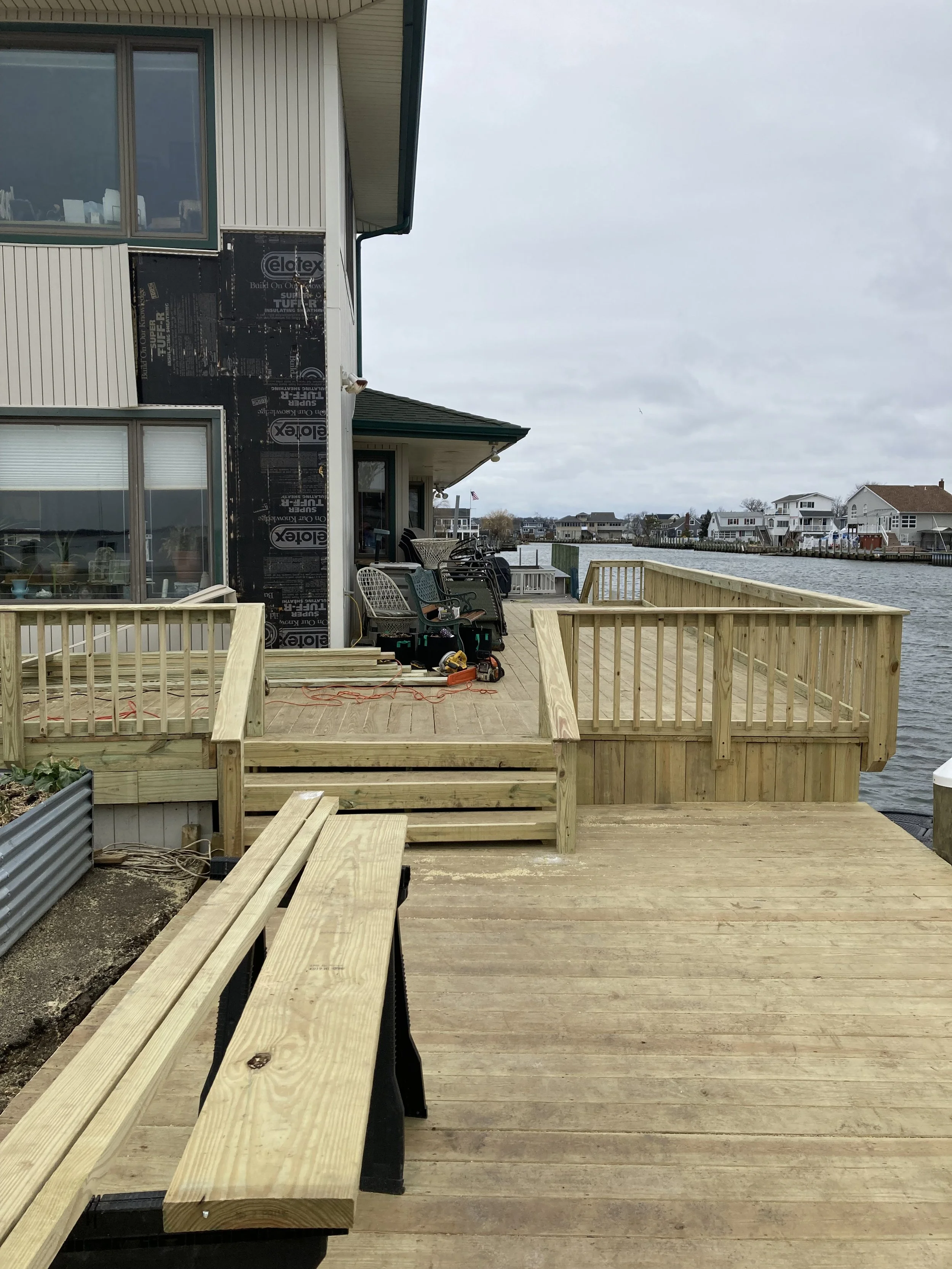 Backyard deck under construction by a waterfront house, with several chairs, some tools, and exposed black house wrap on the siding.