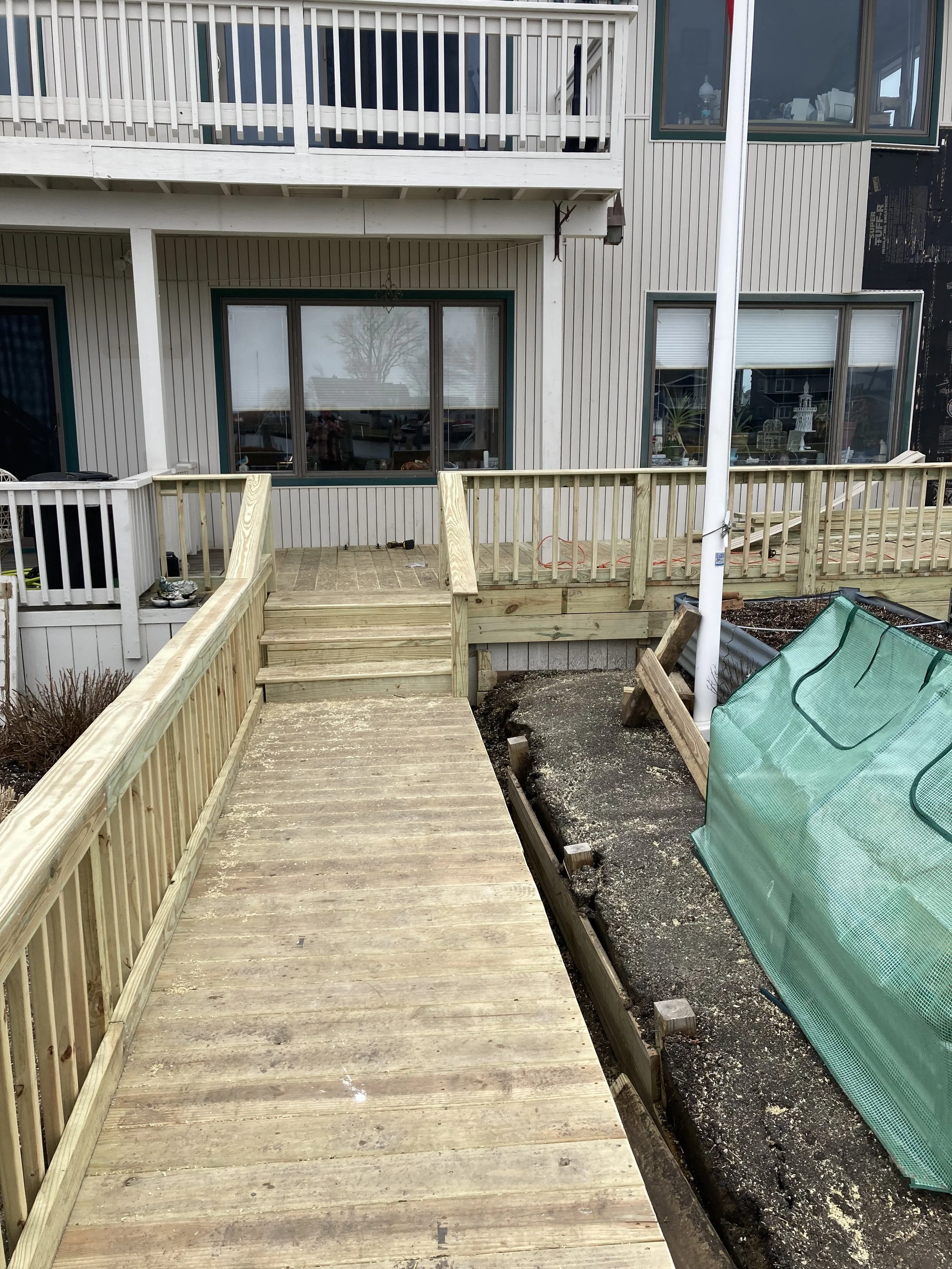 Backyard with wooden ramp and deck under construction, next to a multi-story house with large windows and a balcony.