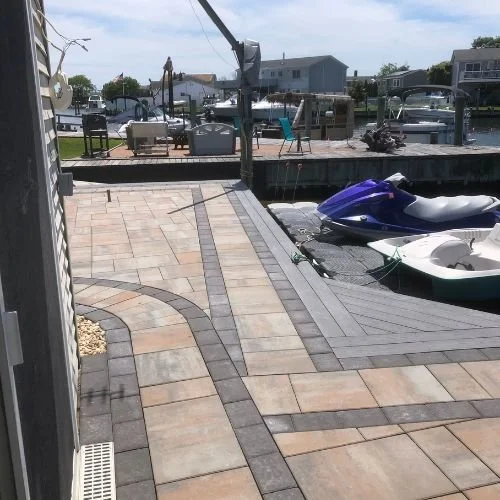 A paved backyard patio with a dock and boats at a marina. The patio has beige and gray tiles with curved patterning, and there is outdoor furniture and a grill in the background.