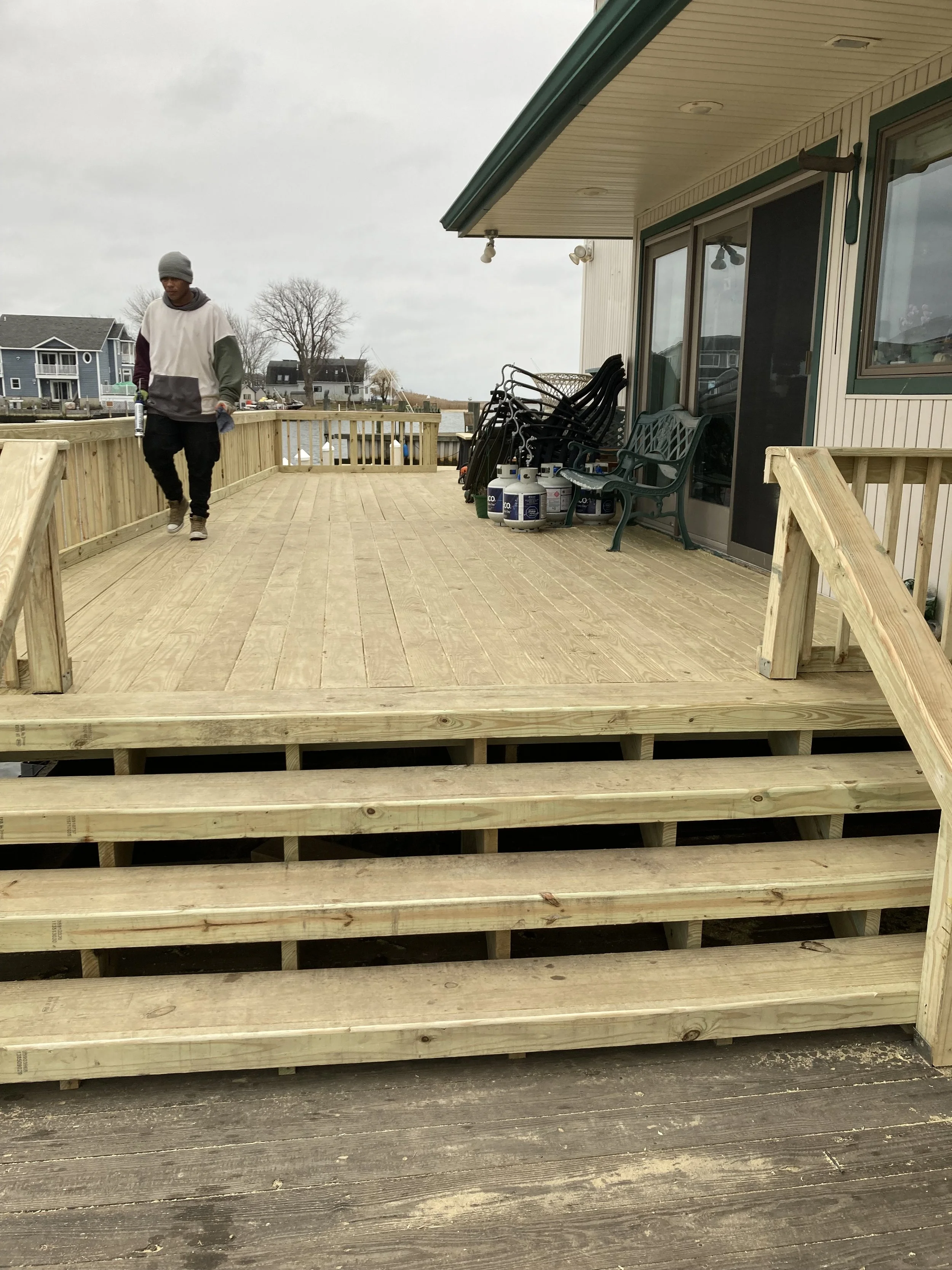 Newly built wooden deck with stairs leading up to it, attached to a house with outdoor furniture and propane tanks, with a person walking on the deck and neighboring houses visible in the background.