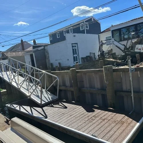 A backyard deck with a metal ramp leading to it, enclosed by a wooden fence. In the background, there is a building with a white exterior and a smaller house with a gray roof. A construction excavator is visible behind the fence, working on the yard, under a blue sky with some clouds.