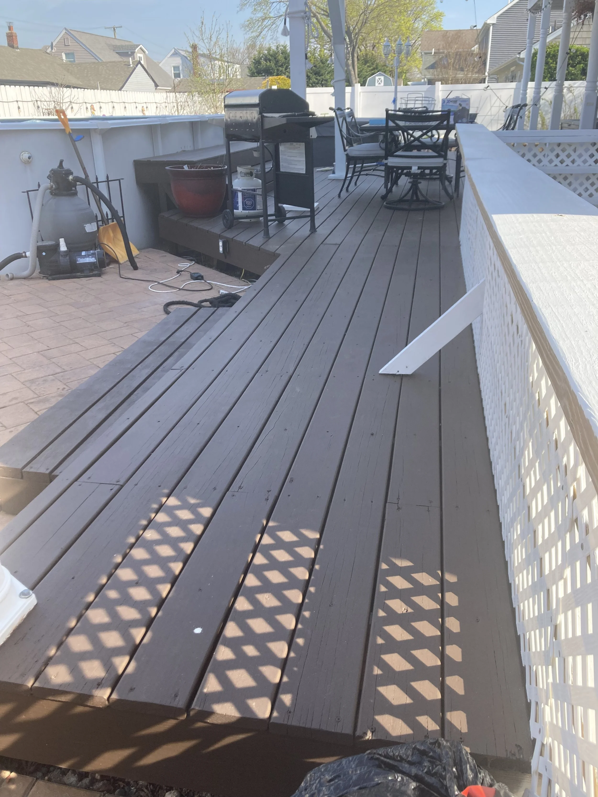 Backyard deck with outdoor furniture, a grill, and pool equipment, surrounded by a white fence and neighboring houses.