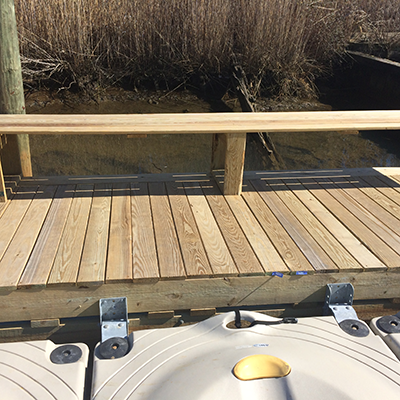 Part of a wooden dock or deck with a railing, overlooking a marshy area with reeds and water, with a boat in the foreground.