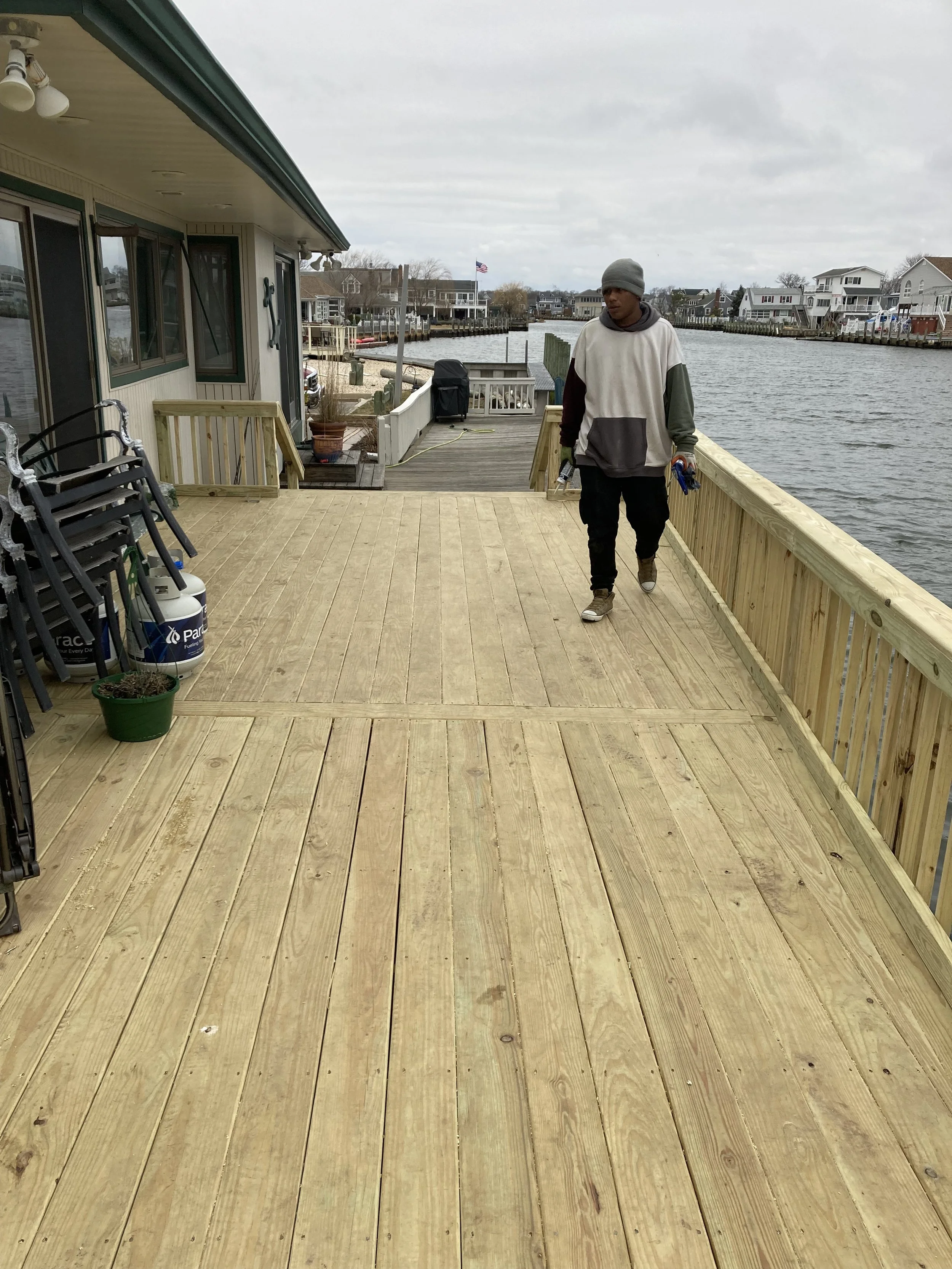 A person walking on a newly built wooden dock beside a house near a waterway, with chairs, a plant, and a grill nearby, and houses across the water in the background.