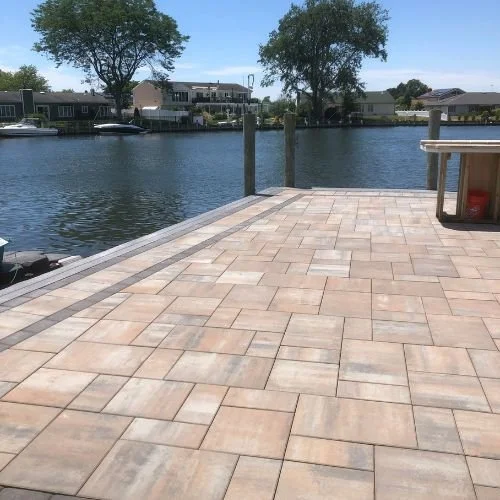 View of a waterfront patio with a paved stone surface, wooden docks, boats, and houses with trees in the background under a clear blue sky.