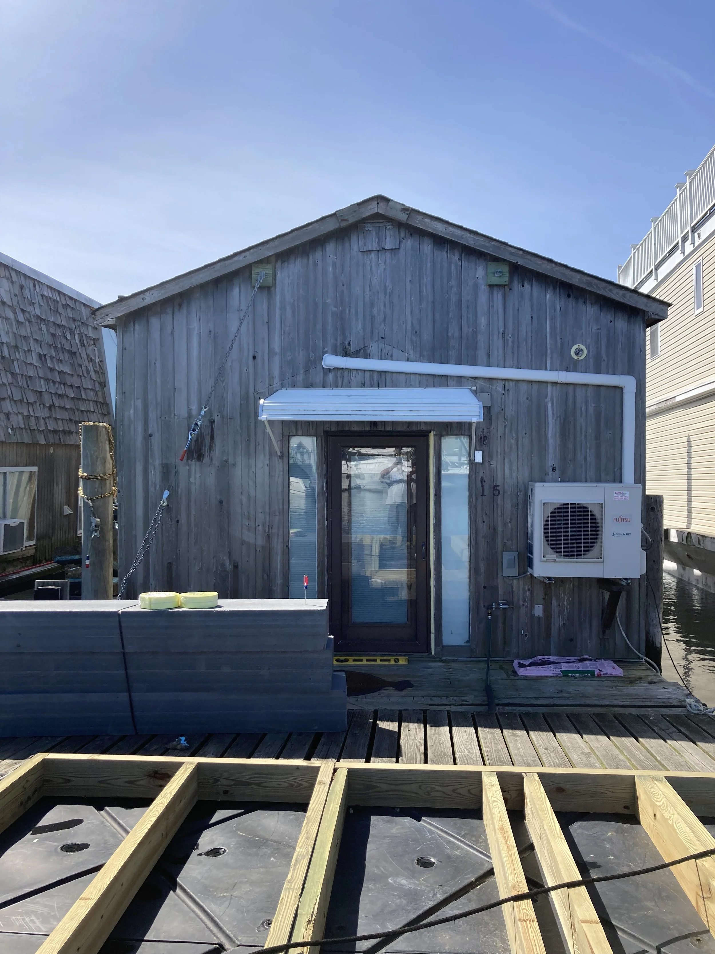 A weathered wooden building under construction, with a door, a window, and an air conditioning unit. Construction materials and tools are visible in the foreground, and part of a deck is being built.
