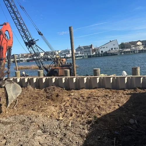 Construction site by water with excavator and wooden pilings in a residential area.