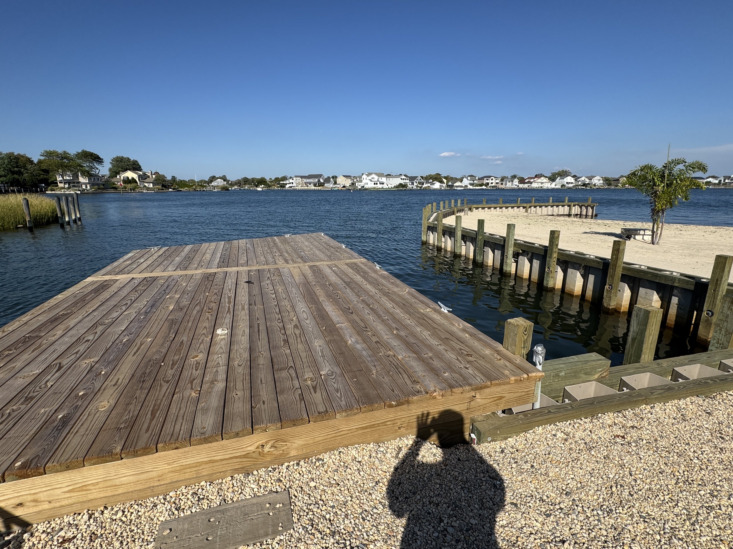 View of a wooden dock extending into a calm body of water with sandy and grassy land on either side, a palm tree on the right, and waterfront houses in the background under a clear blue sky.