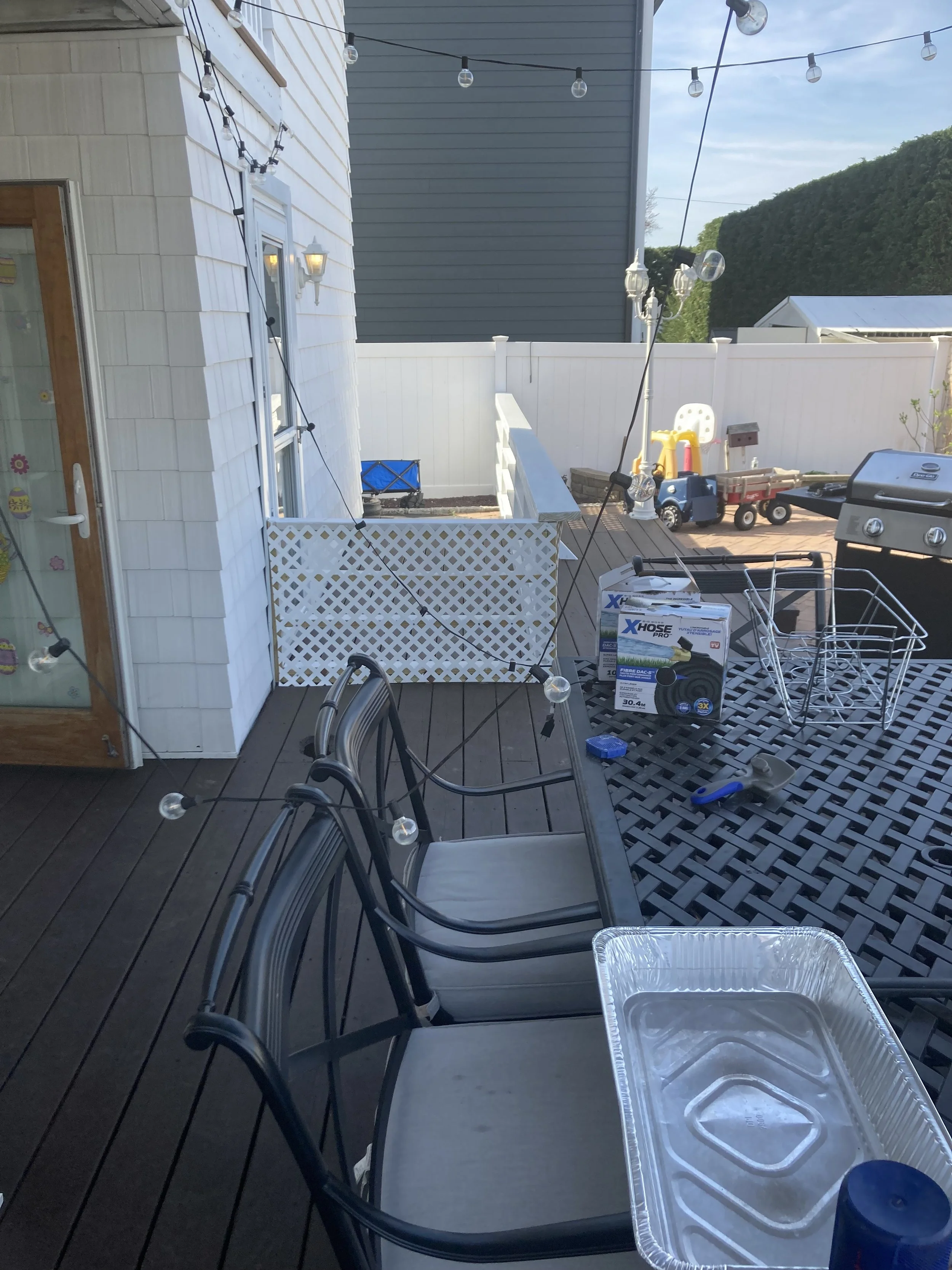 Outdoor deck with chairs, string lights, and various objects including a BBQ grill, toys, a box of garden hose, and a leaf blower.