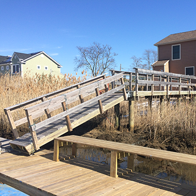 A wooden ramp leading to a floating dock over water, with houses and trees in the background under a clear blue sky.