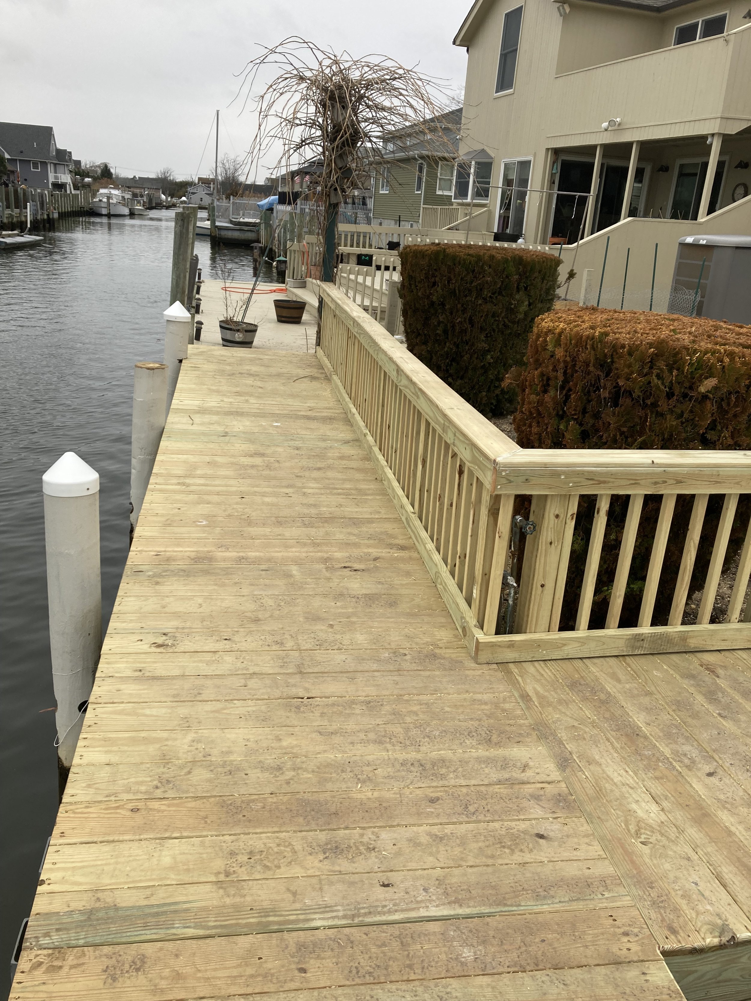 A wooden dock along a residential canal with houses, bushes, and a boat in the distance.