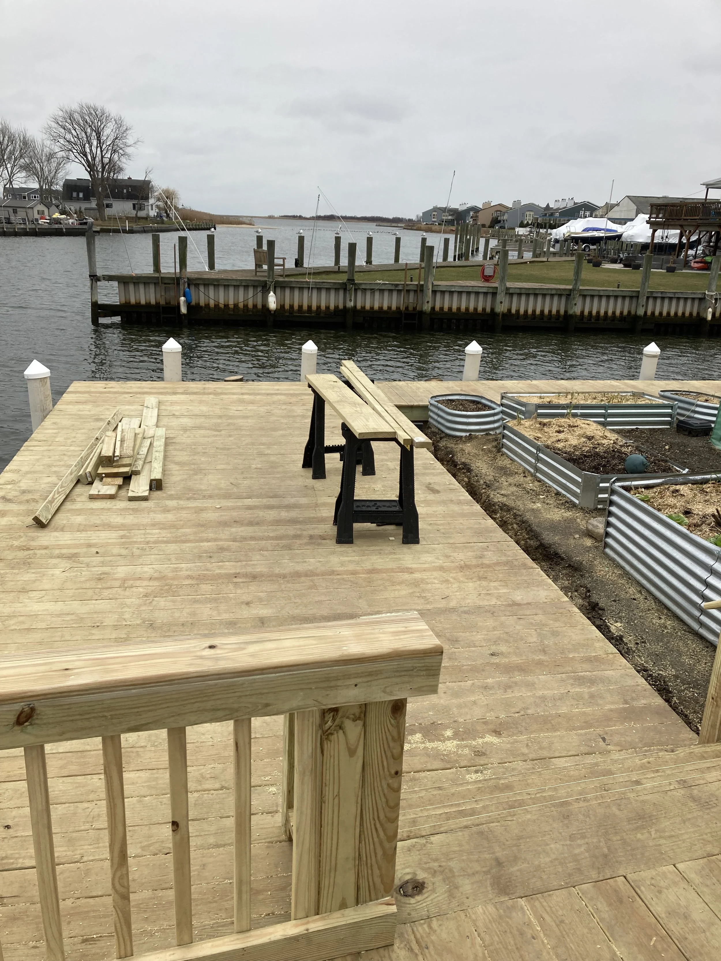 A wooden dock with a bench, some stacked wood planks, and garden beds with soil, overlooking a waterway with other docks, boats, and houses on the shoreline.