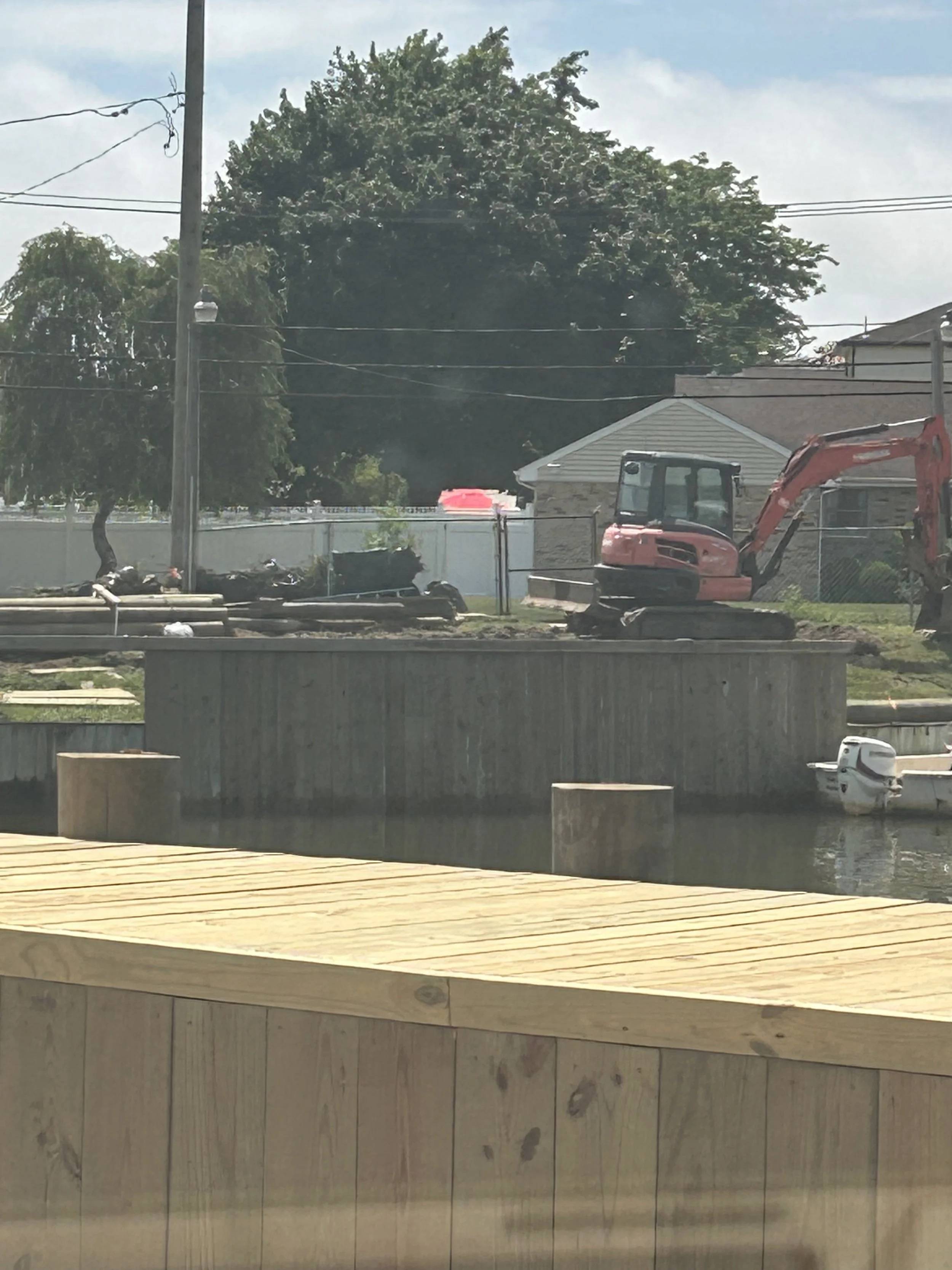 Construction site near a body of water with a small white boat, an orange excavator, and several pipes and wooden planks, with trees and residential houses in the background.