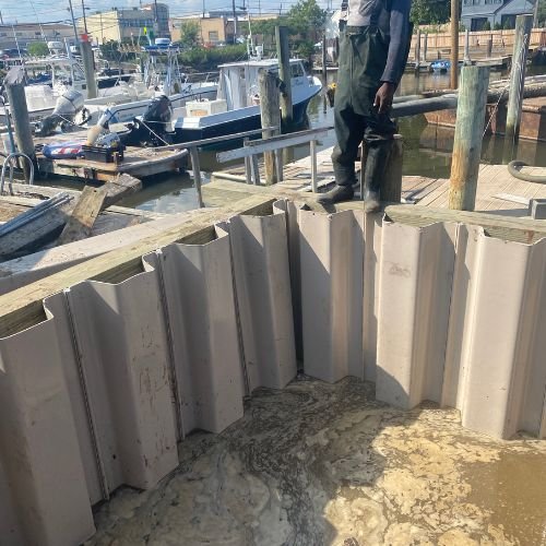 View of a marina with boats docked, and a person standing on a wooden pier near a large, beige, metal storm barrier at a waterfront.
