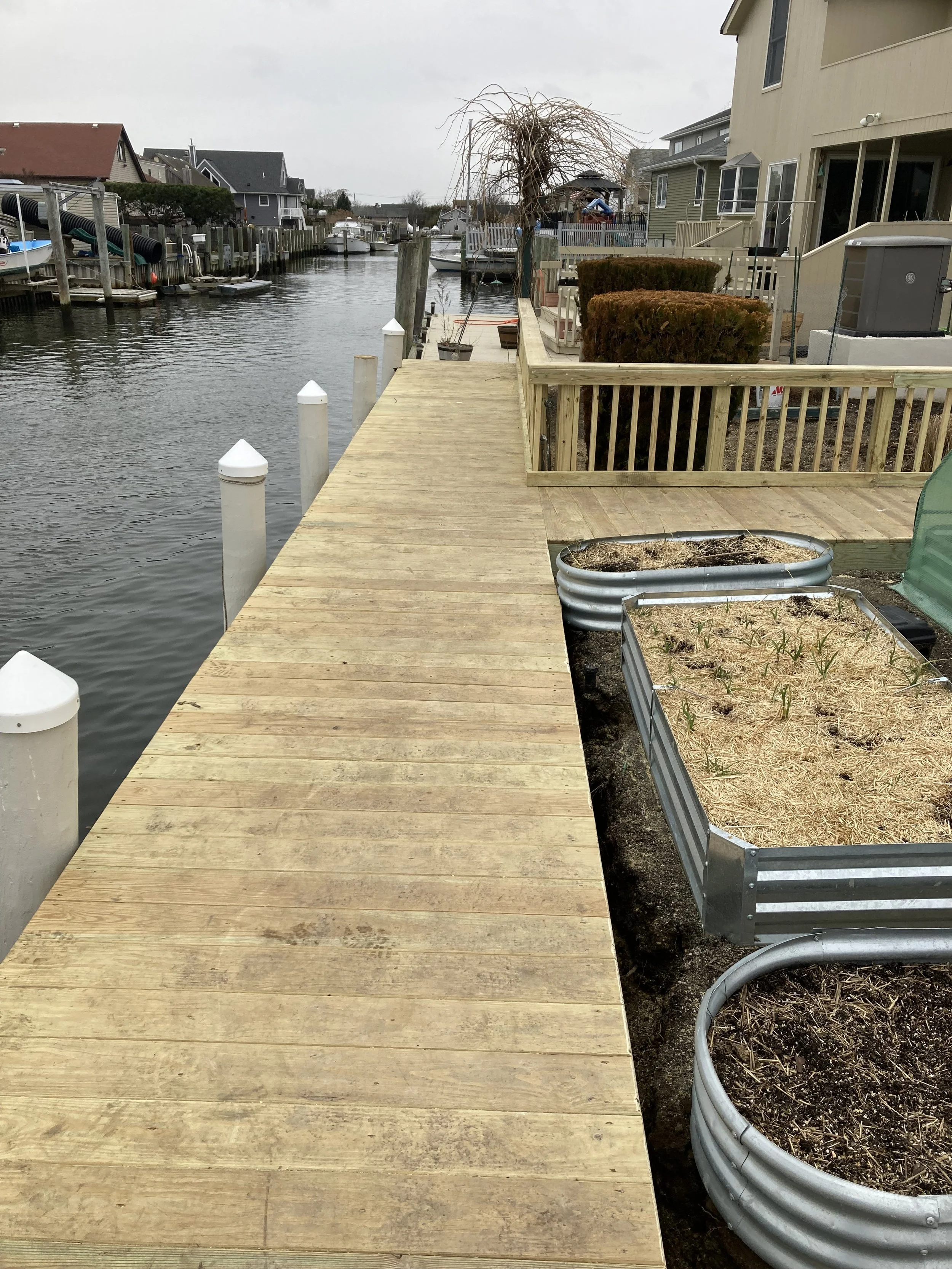 A wooden dock on a canal with houses, boats, and potted plants along the water, under an overcast sky.