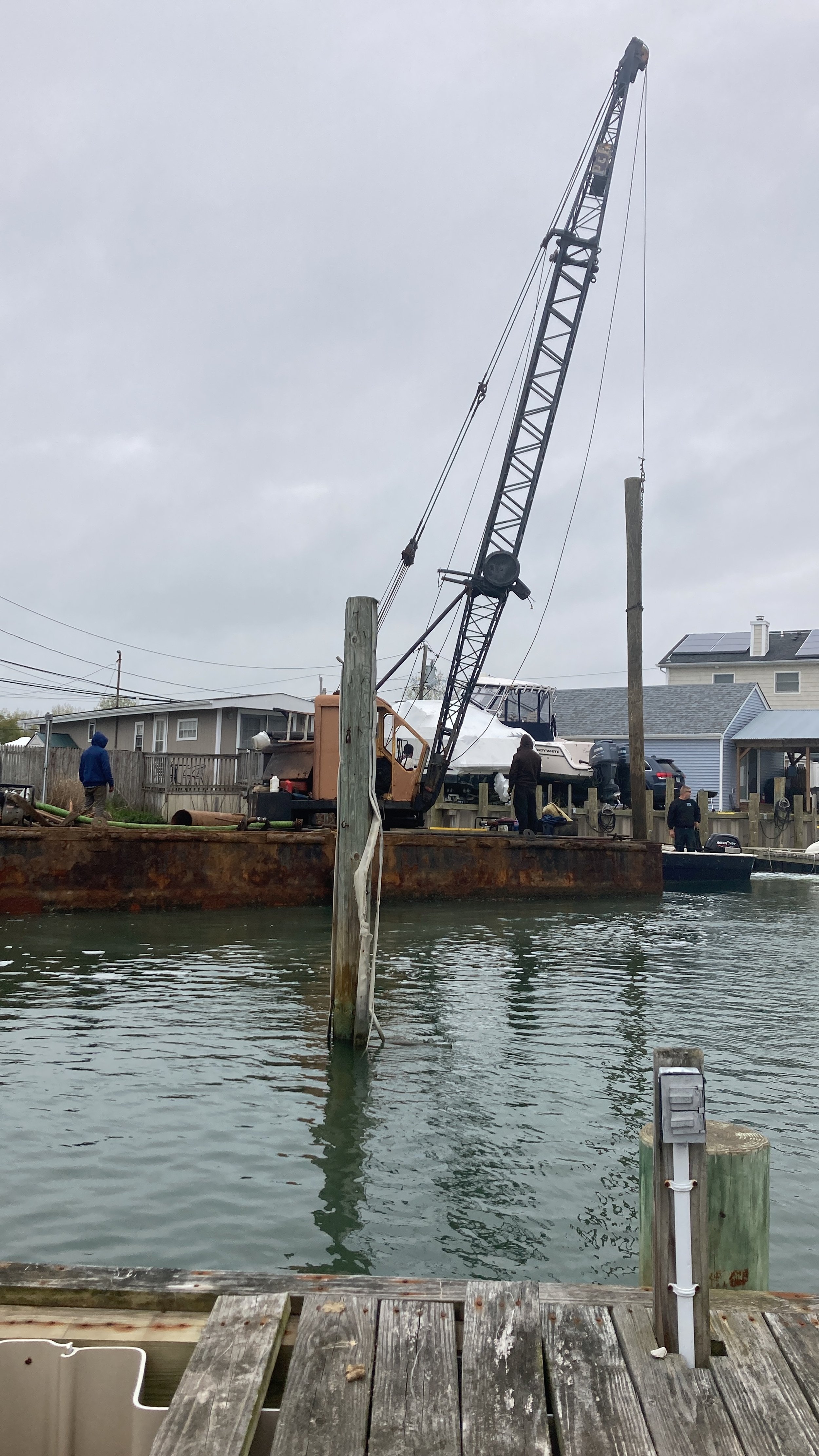 A rusty barge with a crane lifting a boat at a marina on a cloudy day, with residential houses in the background and a person standing nearby.
