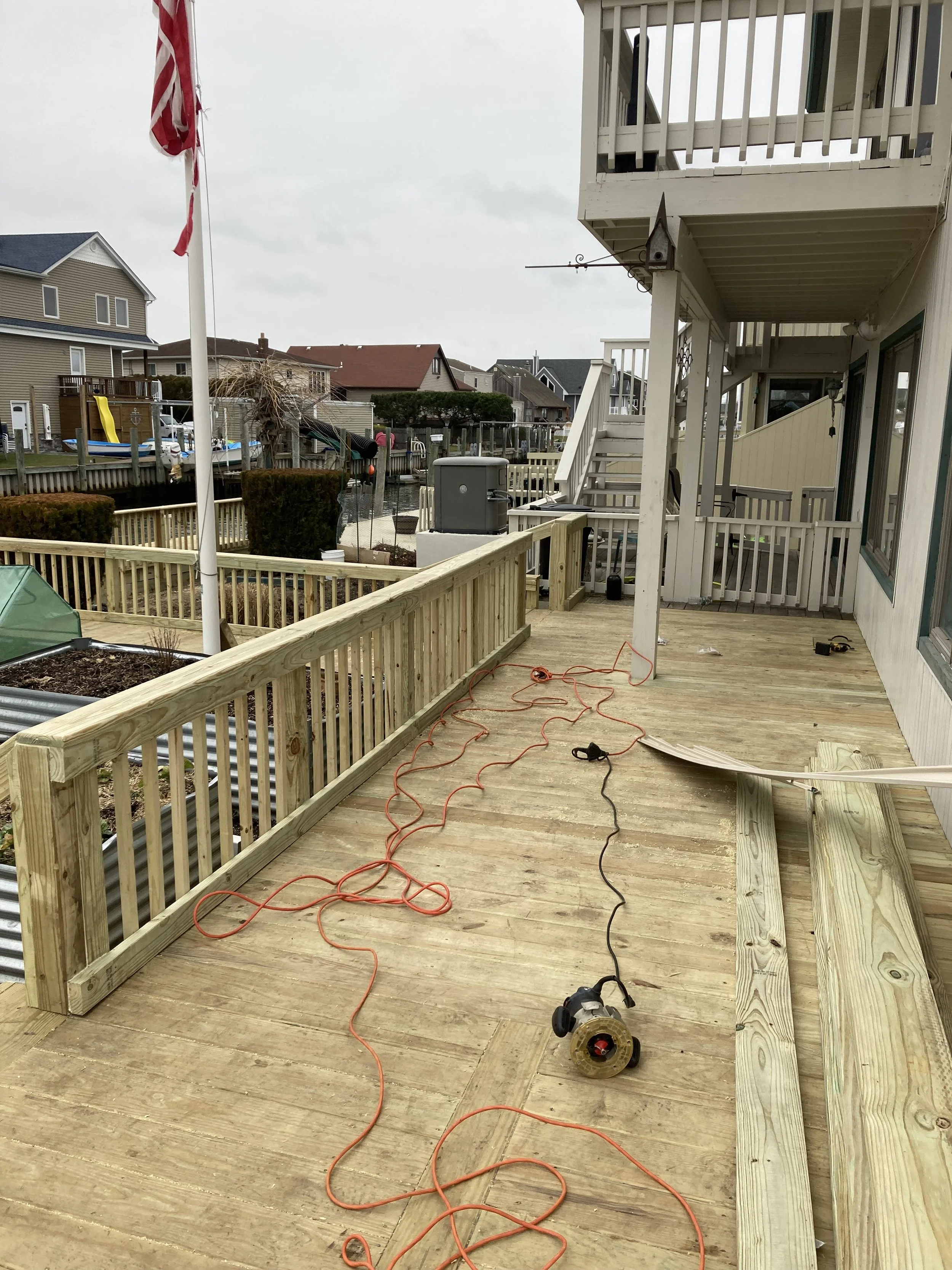 Construction in progress on a wooden deck with tools, extension cords, and a railing, in a residential area with houses and water in the background.