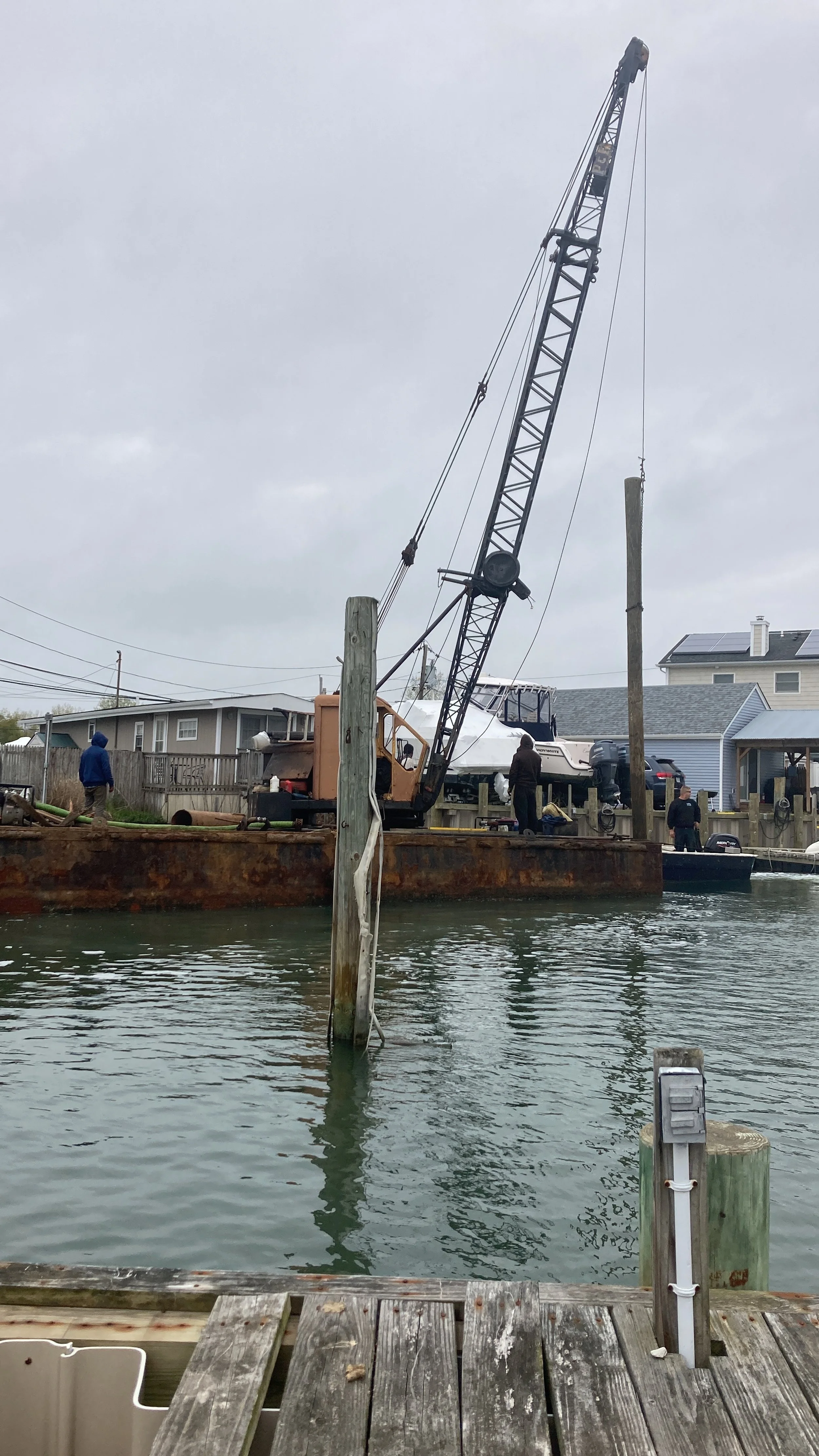 A rusty barge with a crane lifting a boat in a residential marina area under gray, cloudy skies.