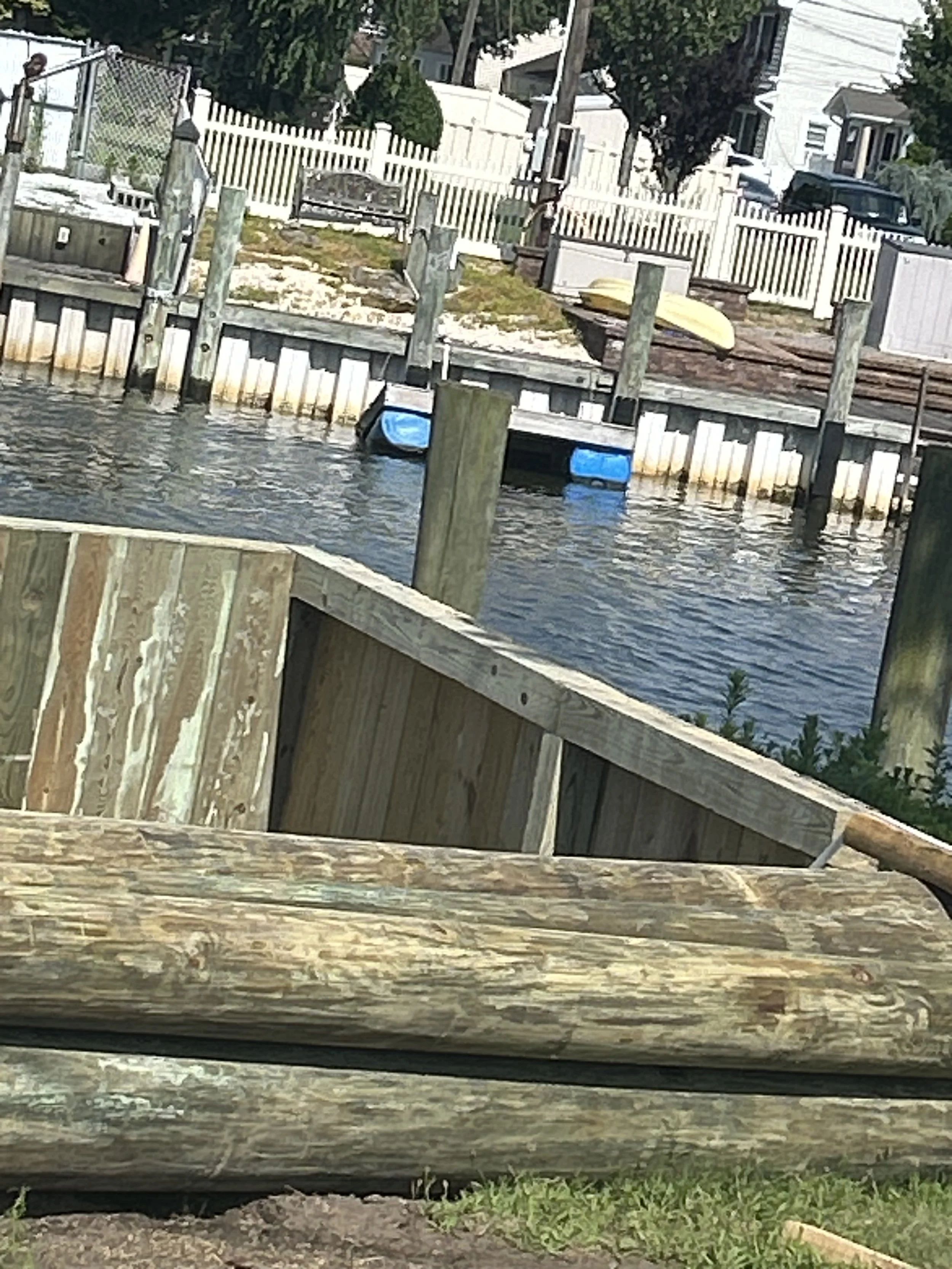 A small dock or boat ramp on a waterway, with weathered wooden railings and pilings, leading to a sandy shoreline with a white picket fence and residential buildings in the background.