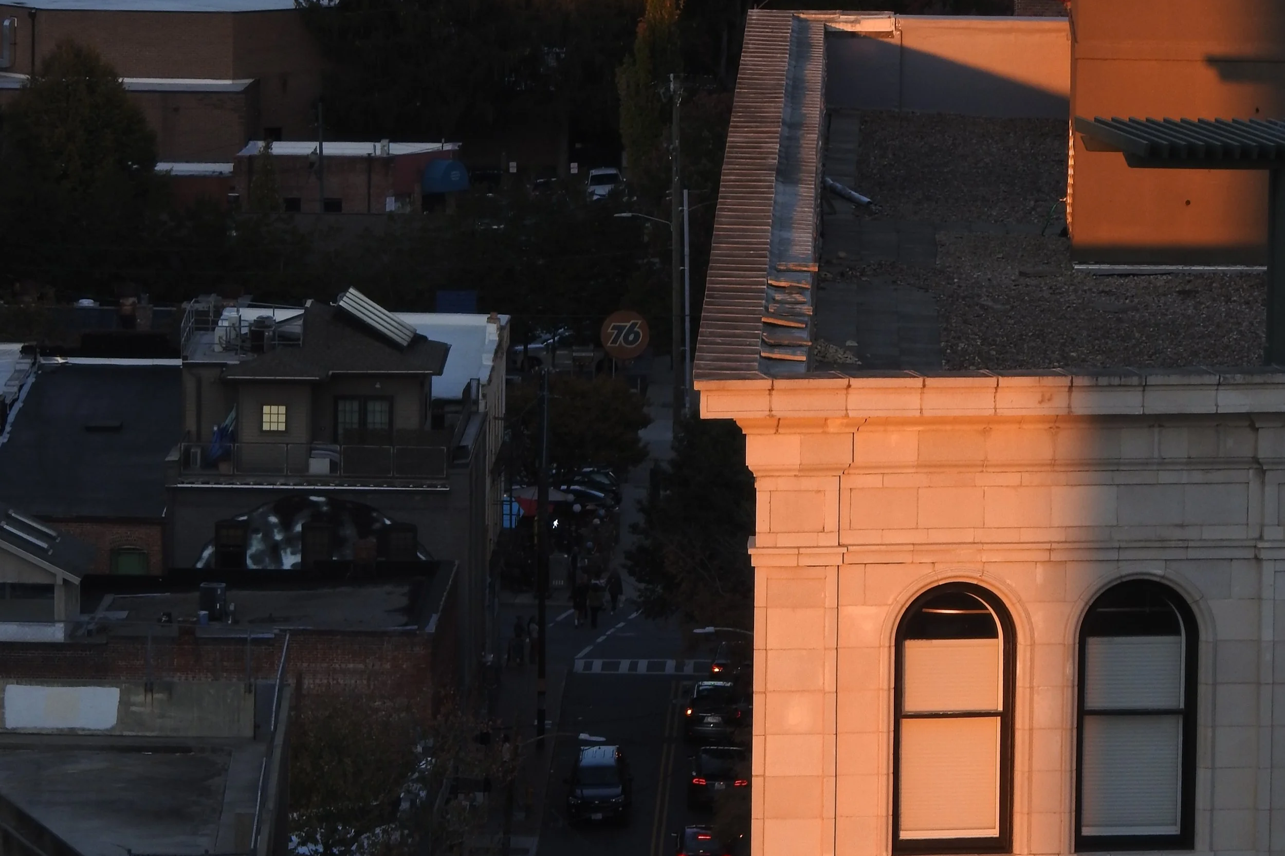 A cityscape view showing the top of a building with a lit orange glow, adjacent rooftops, trees, and a street with parked cars and people walking.