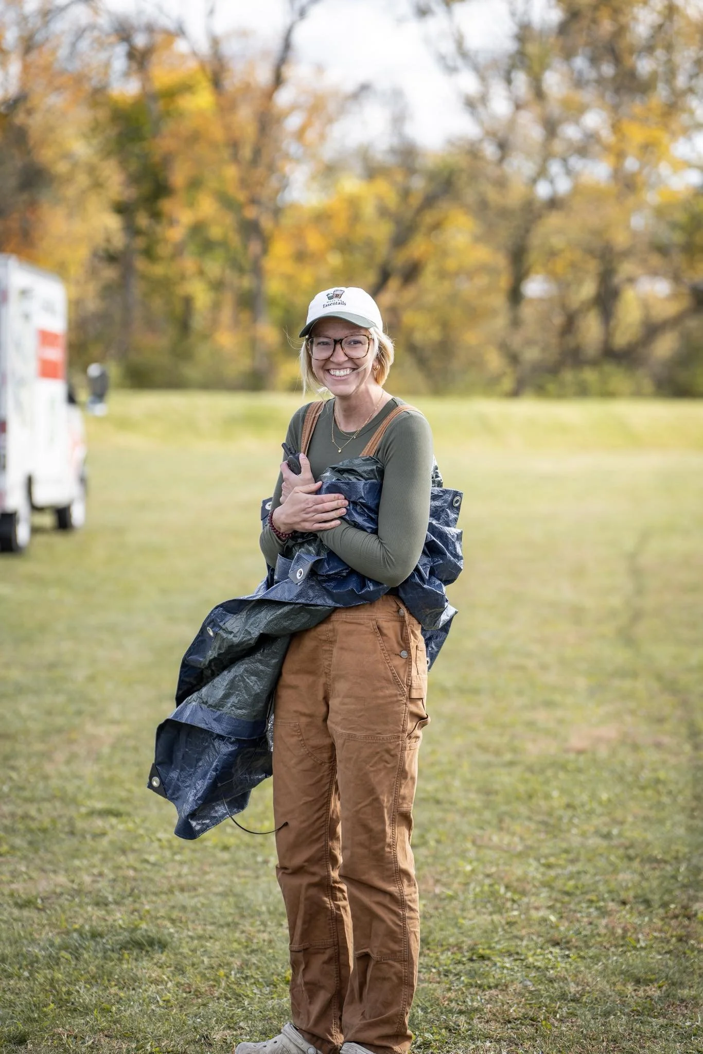 A smiling woman holding a rolled-up tent in an outdoor setting with fall foliage.