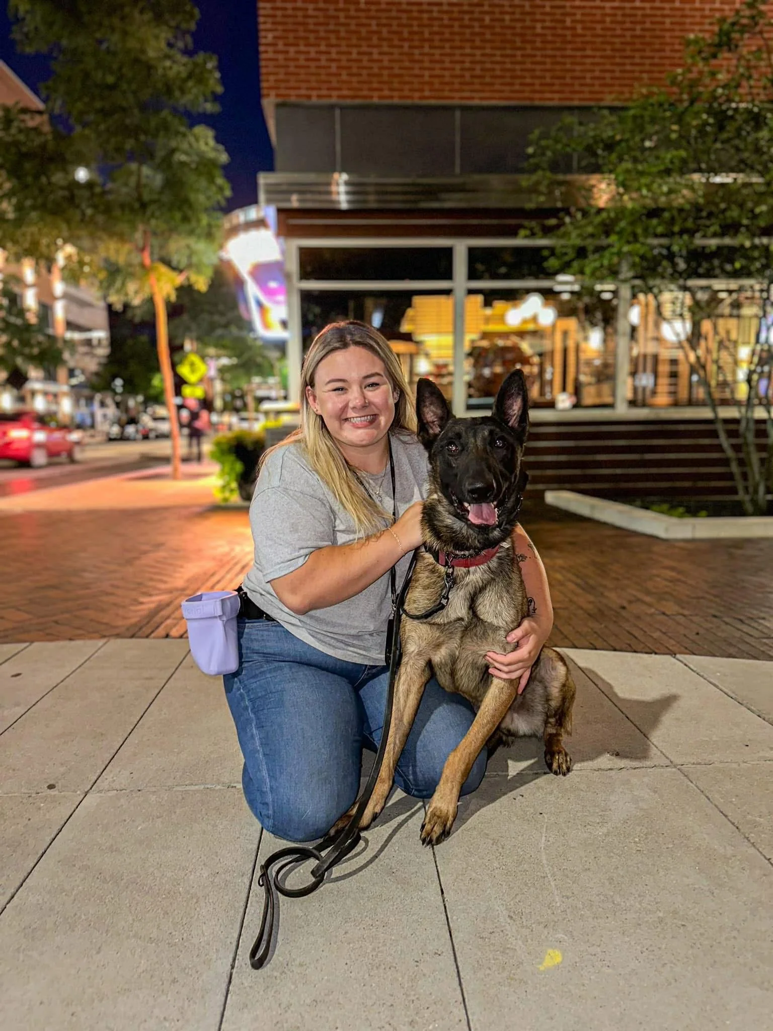 A woman kneeling on the sidewalk, smiling, with a large German Shepherd dog in an urban area at night. The background displays a busy street with cars, trees, and a storefront with illuminated windows.