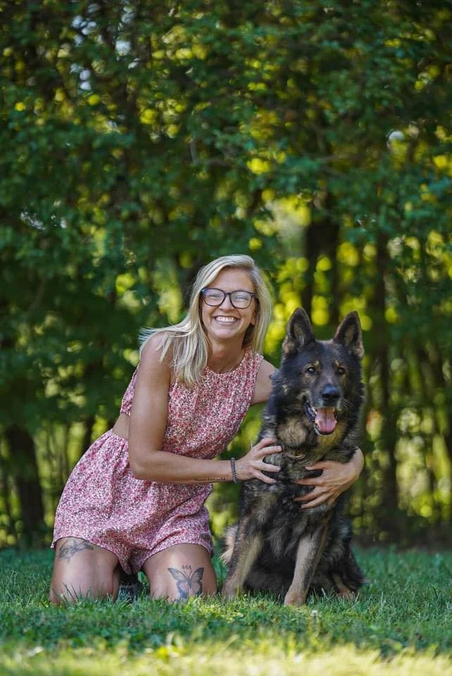 A smiling woman with blonde hair and glasses kneels on the grass next to a large German Shepherd dog outdoors in a wooded area.