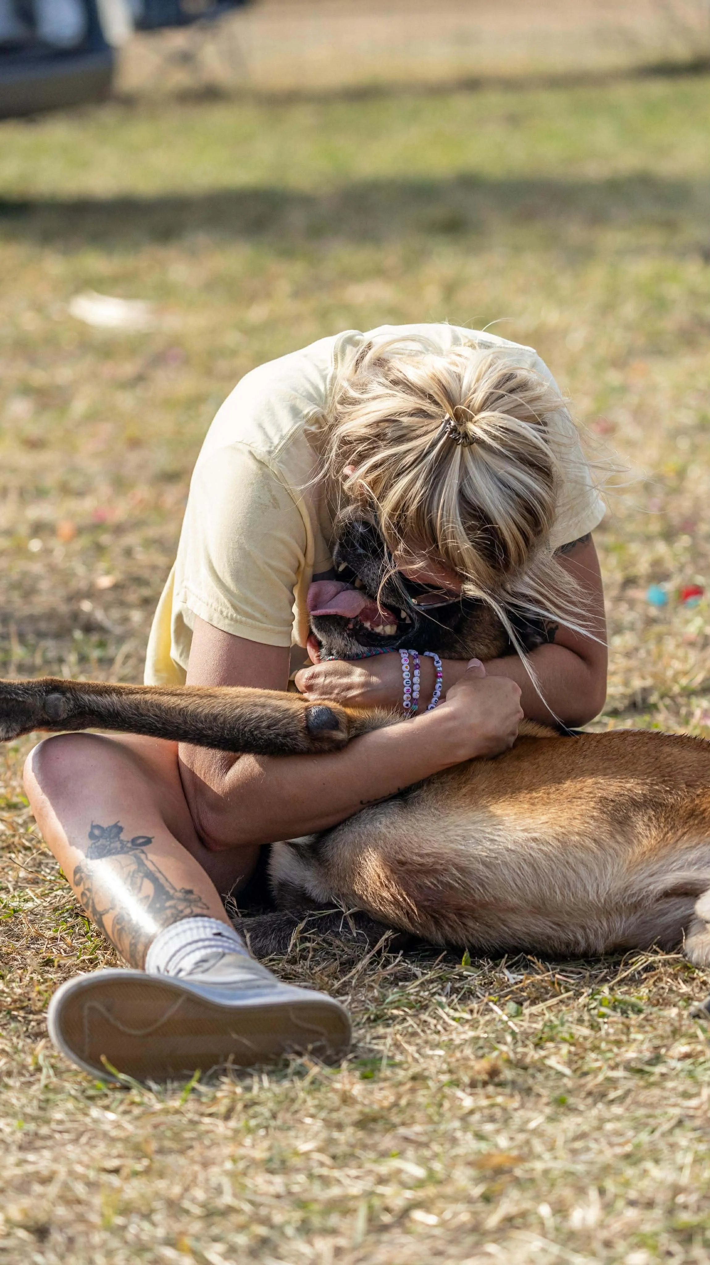 A woman sitting outdoors on the grass, hugging and playing with a German Shepherd dog. She is smiling and appears happy.