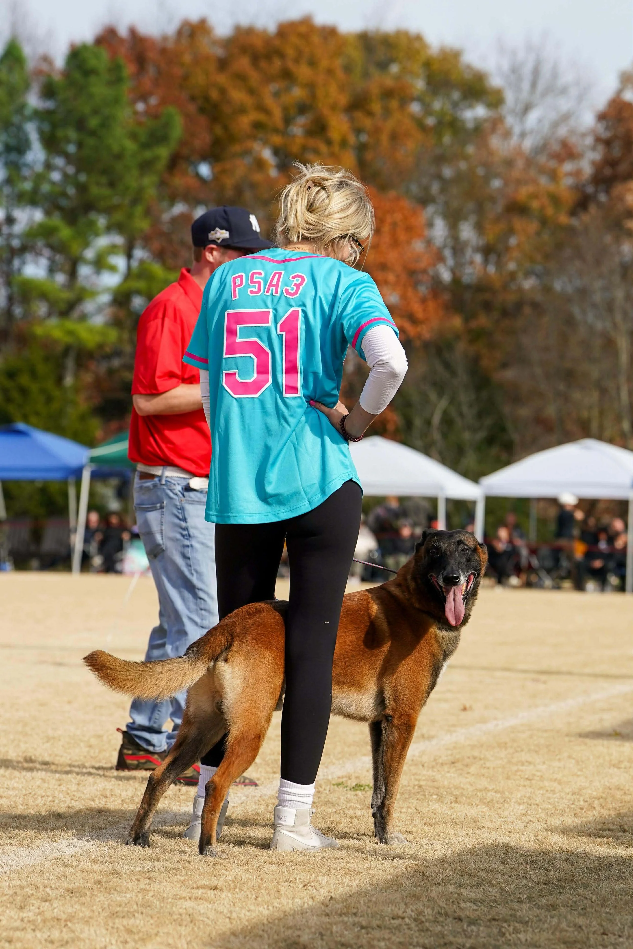 A woman with blonde hair and a turquoise sports jersey with the number 51 stands with her back to the camera on a grassy field, holding a happy brown dog with a black face and a pink tongue hanging out, during an outdoor event with tents and trees wi