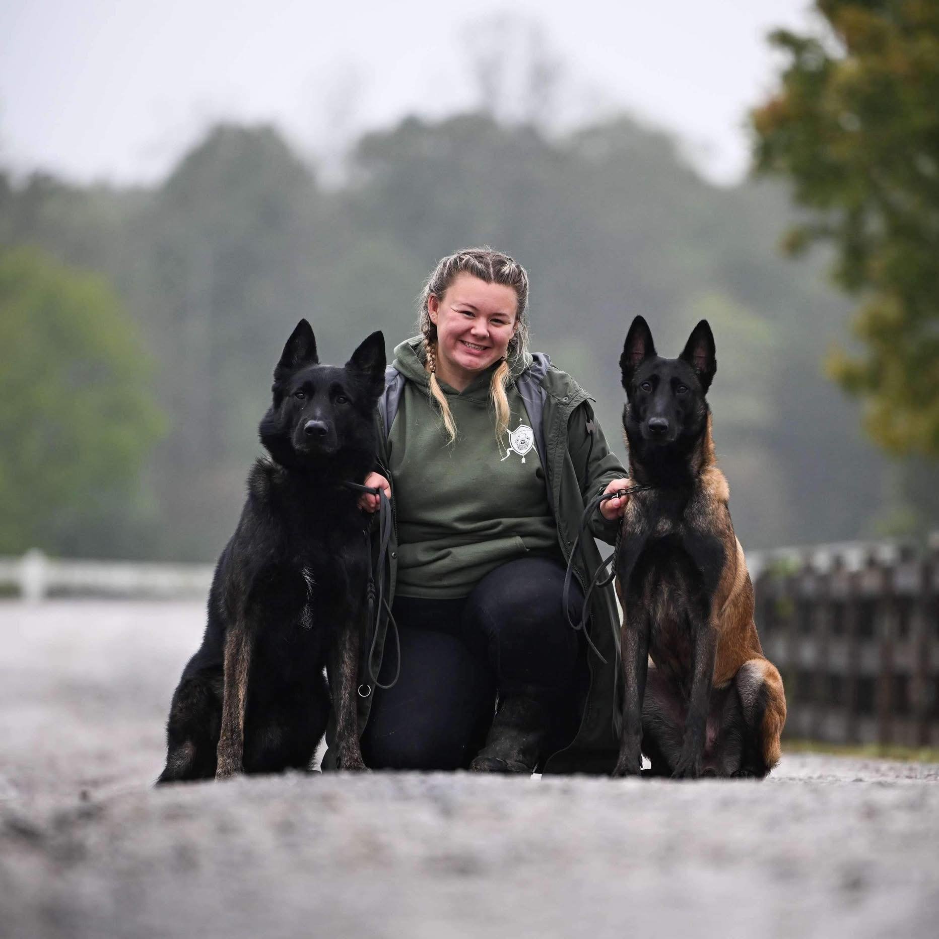 A female police officer kneeling on a bridge, smiling and holding the leashes of two police dogs, one black and one brindle, on a foggy day with trees in the background.