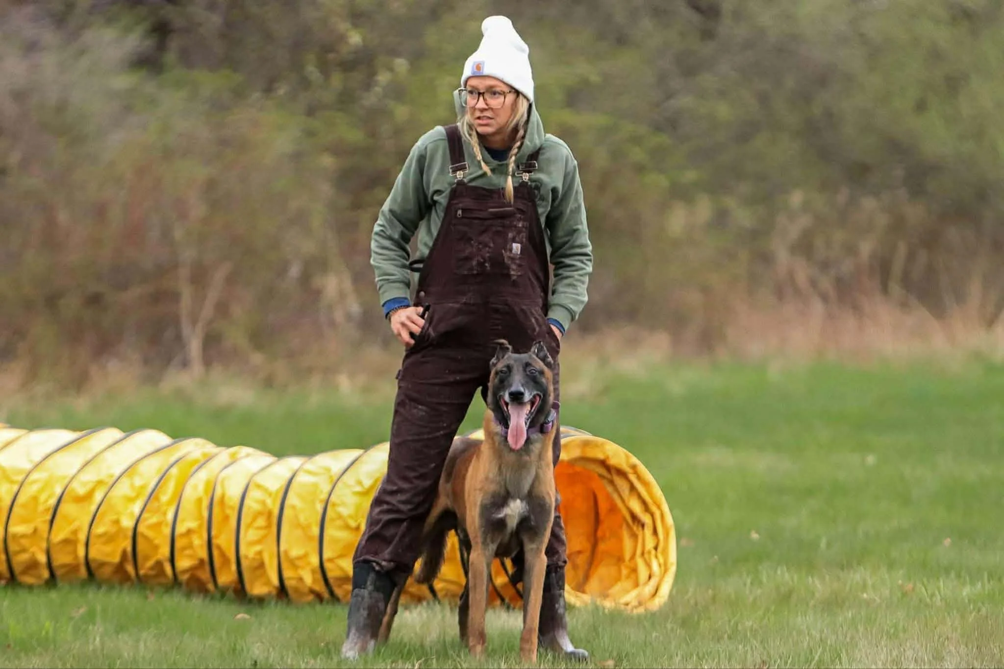 A woman in outdoor gear and a white beanie standing on grass with her tongue out, next to a Belgian Malinois dog in front of a yellow agility tunnel.