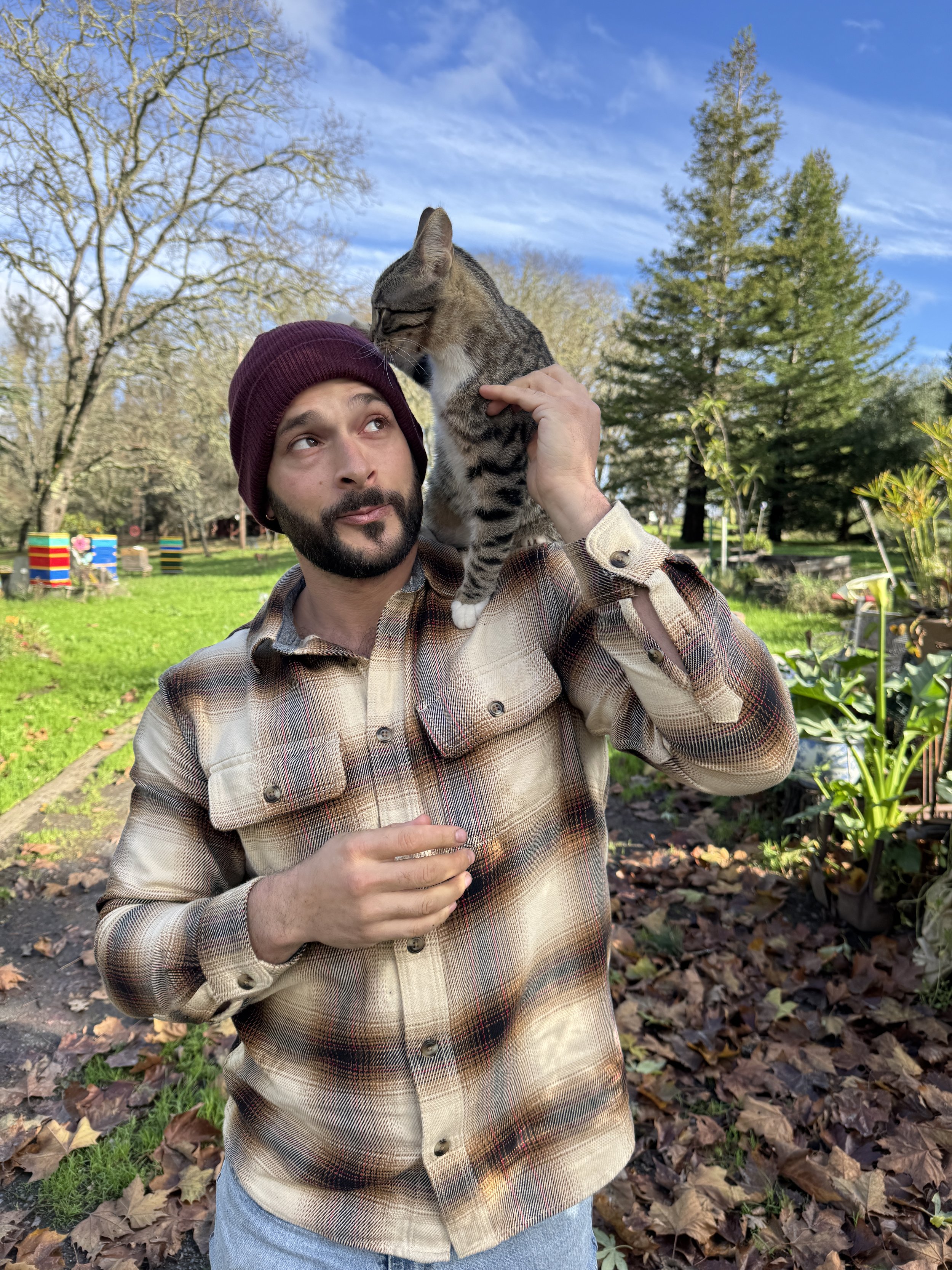 A man wearing a plaid shirt and maroon beanie holding a cat on his shoulder in an outdoor park with trees and grass.