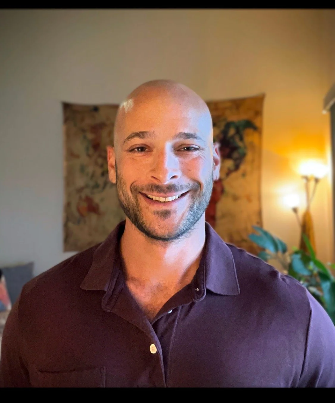 A smiling man with a beard and a shaved head standing indoors, wearing a dark purple shirt with a background of a wall hanging and a lamp.