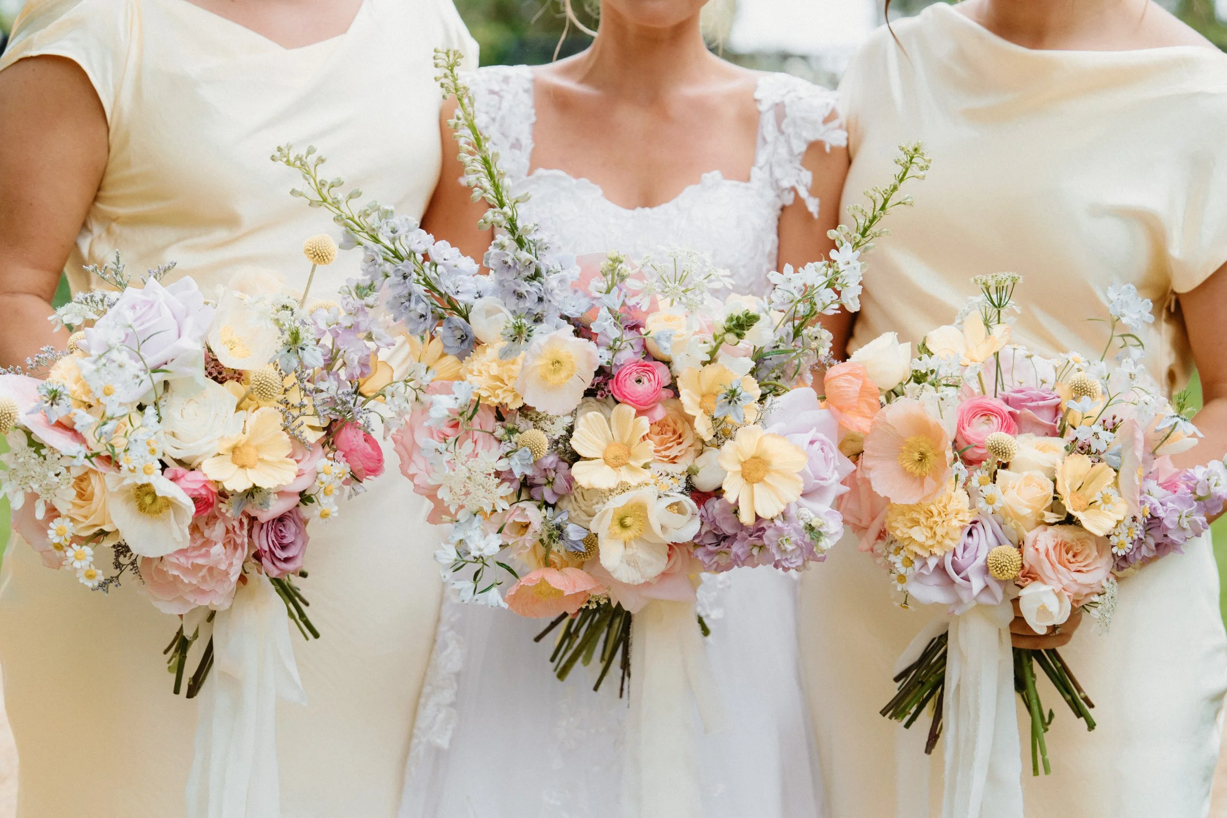 Three women standing together holding large bouquets of pastel-colored flowers, with the woman in the middle wearing a white lace dress and the other two in light yellow dresses.